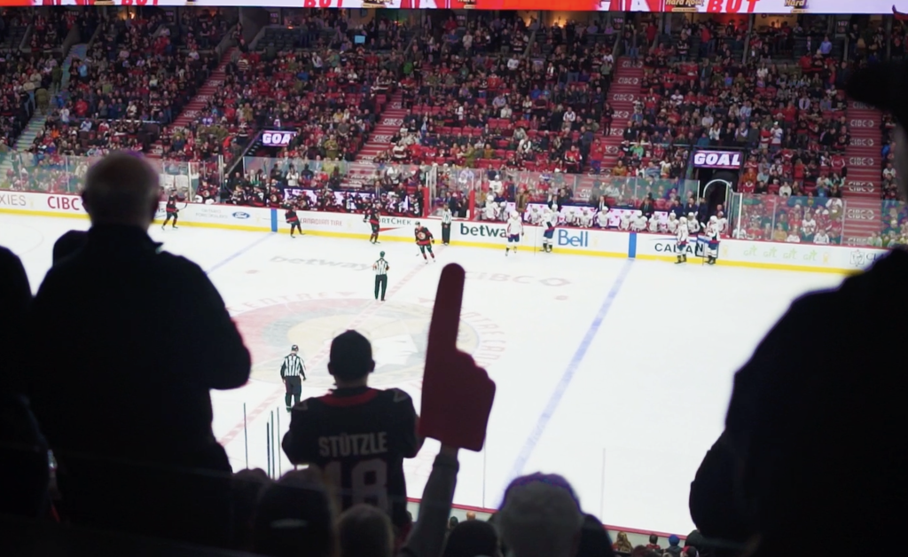 A crowd watches a hockey game in a large arena; a fan in the foreground raises a foam finger while players and officials are on the ice.